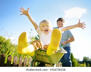 Happy Little Boy Having Fun In A Wheelbarrow Pushing By Dad In Domestic Garden On Warm Sunny Day. Active Outdoors Games For Family With Kids In The Backyard During Harvest Time