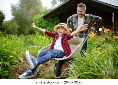 Happy Little Boy Having Fun In A Wheelbarrow Pushing By Dad In Domestic Garden On Warm Sunny Day. Active Outdoors Games For Family With Kids In The Backyard In Summer