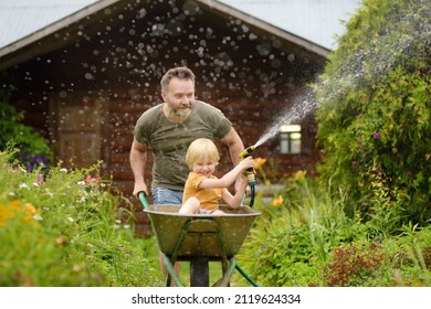 Happy Little Boy Having Fun In A Wheelbarrow Pushing By Dad In Domestic Garden On Warm Sunny Day. Child Watering Plants From A Hose. Active Outdoors Games For Family With Kids In Summer.