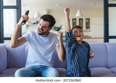 A happy little boy and his cheerful dad are sitting together on the couch in front of the TV, and cheerfully cheering for their favorite football team at home in the living room. - Powered by Shutterstock