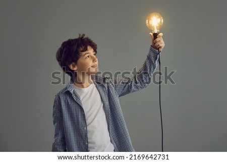 Happy little boy holding shining lit Edison light bulb standing on gray studio background. Smart kid looking at glowing incandescent lightbulb in his hand. Creative idea and inspiration concept