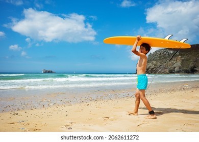 Happy little boy in shorts walk and look at camera hold orange surfboard on the hand to the sea waves on the beach side view