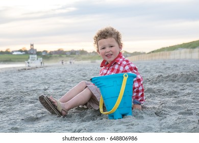 A Happy Little Boy Is Sitting On A New England Beach With A Blue Pail. He Is Wearing A Red And White Checkered Shirt And Has Curly Hair. The Photo Was Shot At The Ocean In Newport, Rhode Island. 