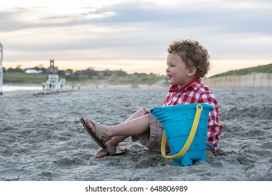 A Happy Little Boy Is Sitting On A New England Beach With A Blue Pail. He Is Wearing A Red  Checkered Shirt And Shorts. The Photo Was Shot In Newport, Rhode Island. He Is Looking Towards The Ocean. 