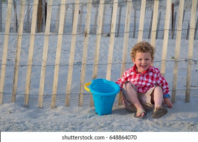 A Happy Little Boy Is Sitting On A New England Beach With A Blue Pail Against A Fence. The Photo Was Shot On A Beach In Newport, Rhode Island. He Is Wearing A Red Shirt, Shorts And Sandals. 