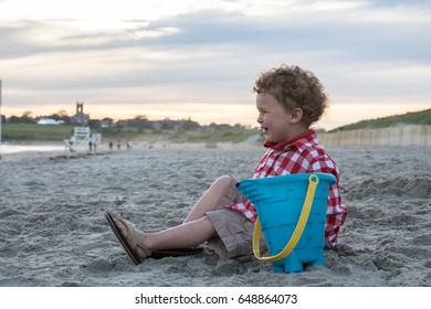 A Happy Little Boy Is Sitting On A New England Beach With A Blue Pail. He Is Wearing A Red  Checkered Shirt And Shorts. The Photo Was Shot In Newport, Rhode Island. He Is Looking Towards The Ocean. 