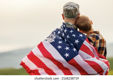 Happy Little Boy Son With American Flag Hugging Father In Military Uniform Came Back From US Army, Rear View Of Male Soldier Reunited With Family While Standing In Green Meadow On July 4th