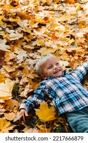 Happy Little Boy In Yellow Bright Coat Throwing Leaves In Sunny Autumn Forest And Jumping Joyfully. Kid Lying In Autumn Leaves. Kid Boy Playing With Leaves In Fall Autumn Park.