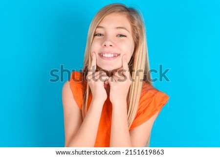 Happy little caucasian kid girl wearing orange T-shirt over blue background with toothy smile, keeps index fingers near mouth, fingers pointing and forcing cheerful smile