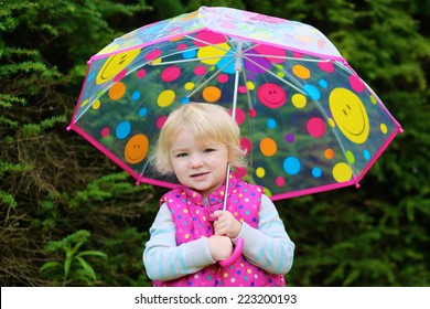 Happy Little Child, Adorable Blonde Curly Toddler Girl Wearing Pink Warm Padded Gilet Holding Colorful Umbrella Playing In The Park On A Sunny Rainy Autumn Day