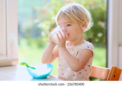 Happy little child, blonde curly toddler girl, enjoying healthy breakfast eating oatmeal porridge and drinking milk sitting in high chair at bright sunny kitchen next to big garden view window - Powered by Shutterstock