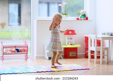 Happy Little Child, Cute Blonde Toddler Girl Having Fun Dancing Indoors In A Sunny White Room At Home Or Kindergarten