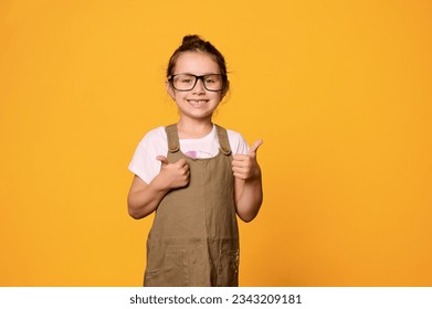 Happy little child girl in eyeglasses, gestures with thumbs up, smiles with a cheerful toothy smile, looking at camera, isolated over orange studio background. Smart pupil. Teacher's Day. School girl - Powered by Shutterstock