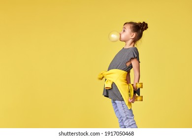 Happy Little Child Girl With Yellow Skateboard Blowing A Big Bubble Gum Over Yellow Background. Copy Space