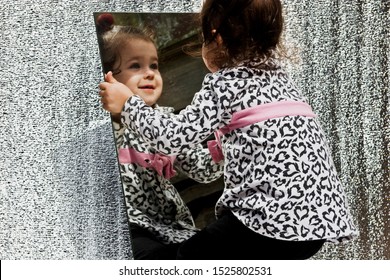 Happy Little Child Grimaces In Front Of A Mirror. A Little Girl Looks At Her Reflection In The Mirror Close-up.