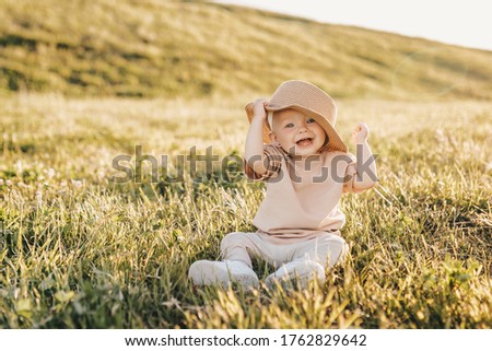 A happy little child laughs poses and plays with a hat while sitting on the grass in a summer park at sunset