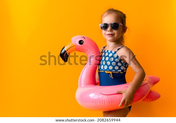 happy little child swimmer sport girl wear sun\
glasses swimsuit, Kid Inflatable swimming ring Pink Flamingo point\
to side away, isolated orange background. childhood, hot summer\
vacation hobby concept