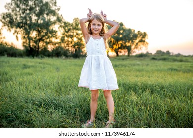 Happy Little Child In White Dress Making Bunny Ears With Fingers On Sunset