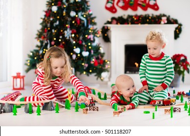 Happy Little Children In Matching Pajamas Playing With Christmas Presents - Wooden Toy Railroad And Car. Family Xmas Morning In Decorated Living Room With Kids Gifts, Fireplace And Christmas Tree. 