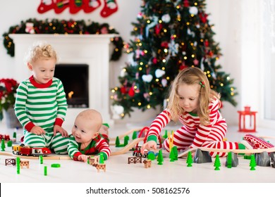 Happy Little Children In Matching Pajamas Playing With Christmas Presents - Wooden Toy Railroad And Car. Family Xmas Morning In Decorated Living Room With Kids Gifts, Fireplace And Christmas Tree. 