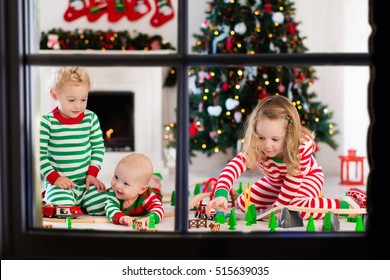 Happy Little Children In Matching Pajamas Playing With Christmas Presents - Wooden Toy Railroad And Car. Family Xmas Morning In Decorated Living Room With Kids Gifts, Fireplace And Christmas Tree. 