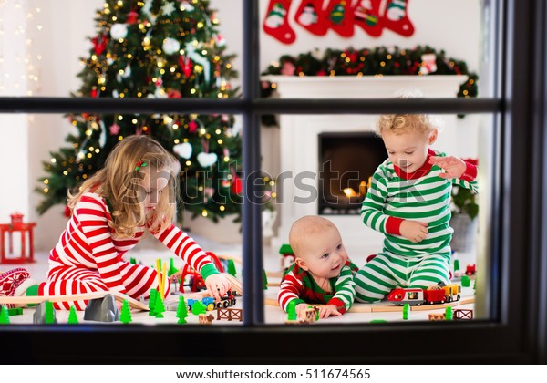 Happy little children in matching pajamas playing with
Christmas presents - wooden toy railroad and car. Family Xmas
morning in decorated living room with kids gifts, fireplace and
Christmas tree. 