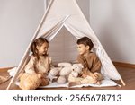 Happy little children siblings boy and girl sitting in a wigwam tent, playing and having fun at home. Children