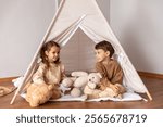 Happy little children siblings boy and girl sitting in a wigwam tent, playing and having fun at home. Children