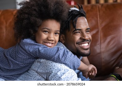 Happy Little Cute African American Child Girl Embracing Young Brazilian Father, Resting Together On Cozy Sofa, Enjoying Stress Free Weekend Time At Home, Good Family Relations Concept.