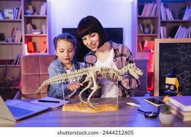 Happy little daughter and her mother sitting together at table and study fossil animals with magnifying glass at evening time. Two Caucasian women wearing casual clothes making prehistorical research. - Powered by Shutterstock