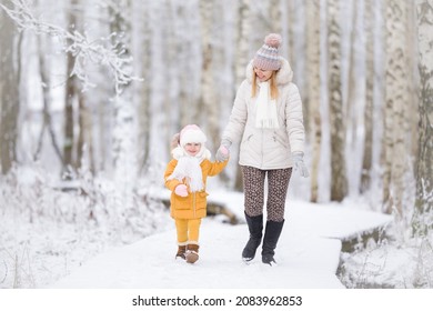 Happy little daughter and young adult mother walking on white snow covered trail at birch tree forest. Spending time together in beautiful cold winter day. Enjoying stroll after blizzard. Front view.