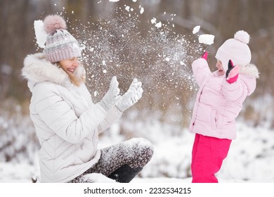 Happy little daughter and young adult mother throwing fresh white snow at park. Lovely fun moment. Spending time together and enjoying cold winter day. Side view.