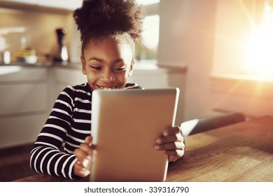 Happy little ethnic black girl sitting at home in the kitchen reading on a tablet computer grinning with pleasure, bright sun glow behind