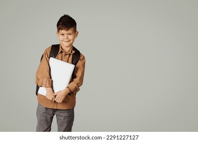 Happy little european child 6 years old pupil in casual with backpack and laptop isolated on gray background, studio. Childhood, knowledge, education and study in elementary school with device