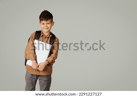 Happy little european child 6 years old pupil in casual with backpack and laptop isolated on gray background, studio. Childhood, knowledge, education and study in elementary school with device