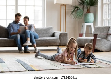 Happy Little Friendly Children Siblings Lying On Floor Carpet Involved In Drawing Pictures In Paper Album While Smiling Young Parents Relaxing On Sofa Using Digital Computer Tablet In Living Room..