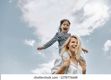 Happy Little Funny Daughter On A Piggy Back Ride With Her Happy Mother On The Sky Background. Loving Woman And Her Little Girl Playing In The Park. Mom And Kid Has Fun Outside. Mother's Day