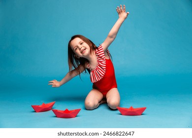 A happy little funny girl in red swimsuit playing with paper boats on blue background. A little girl surrounded by colorful paper boats. The day of the Navy. Adventure concept. Travel time - Powered by Shutterstock