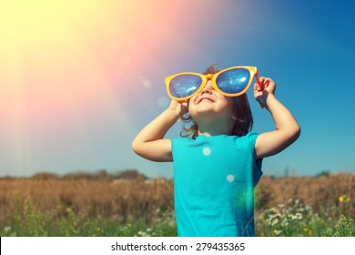Happy little girl with big sunglasses walking in the meadow and looking at the sun