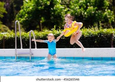 Happy Little Girl And Boy Holding Hands Jumping Into Outdoor Swimming Pool In A Tropical Resort During Family Summer Vacation. Kids Learning To Swim. Focus On Boy.