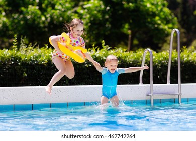 Happy Little Girl And Boy Holding Hands Jumping Into Outdoor Swimming Pool In A Tropical Resort During Family Summer Vacation. Kids Learning To Swim. Focus On Boy.