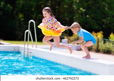 Happy Little Girl And Boy Holding Hands Jumping Into Outdoor Swimming Pool In A Tropical Resort During Family Summer Vacation. Kids Learning To Swim. Water Fun For Children.