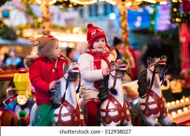 Happy Little Girl And Boy In Warm Jacket And Red Knitted Hat And Scarf Riding Carousel Horse During Family Trip To Traditional German Christmas Market. Kids At Xmas Outdoor Fair On Snowy Winter Day.