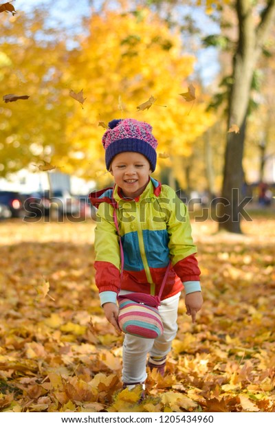 Happy little girl in bright clothes playing\
with leaves in city park in the\
autumn