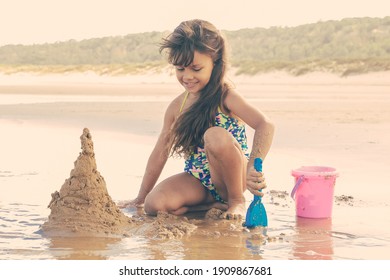 Happy Little Girl Building Sandcastle With Plastic Shovel, Sitting On Wet Sand By Water, Enjoying Vacation On Beach By Ocean. Summer Holidays Or Childhood Concept