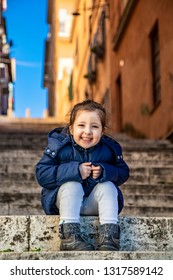 An happy little girl (Caucasian) smiles and enjoys sitting on a step of a staircase, wearing a blue coat, on a sunny winter day. Glimpse of blue sky between the buildings. Frascati, Rome, Lazio, Italy - Shutterstock ID 1317589142