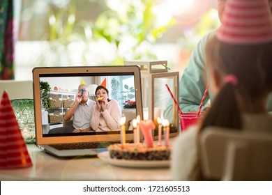 Fröhliches kleines Mädchen, das zu Hause Geburtstag feiert, mit Eltern und großen Eltern auf Videoanruf. Laptop mit Seniorenpaar online, Kuchen mit Kerzen auf dem Tisch.