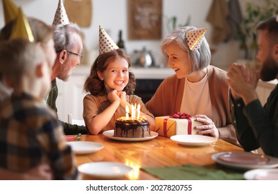 Happy little girl celebrating birthday with family at home, looking at cake with lit candles, making wish while sitting at table in kitchen with smiling loving parents, grandparents and small brother