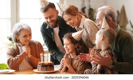 Happy little girl celebrating birthday with family at home, looking at cake with lit candles, making wish while sitting at table in kitchen with smiling loving parents, grandparents and small brother - Powered by Shutterstock