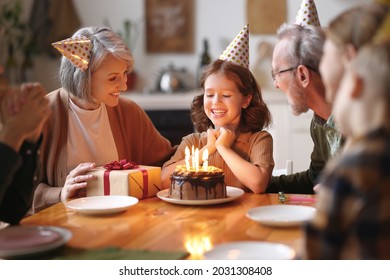 Happy little girl celebrating birthday with family at home, looking at cake with lit candles, making wish while sitting at table in kitchen with smiling loving parents, grandparents and small brother - Powered by Shutterstock